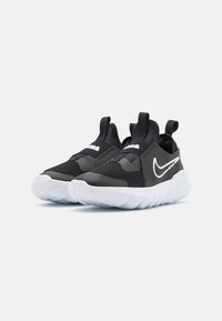 Zapatillas deportivas negras con un diseño sin cordones, parte superior de malla texturizada, cuello acolchado y suela de goma blanca con un logo prominente.