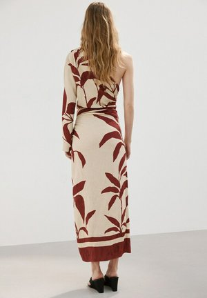 Kobieta o długich włosach ubrana w kremowo-rdzawą, asymetryczną sukienkę z długim krojem i liściastym wzorem oraz czarne koturny, stojąca tyłem.