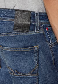 Jeans in denim blu scuro con etichetta in pelle nera con testo in rilievo sulla fascia della vita, cuciture dorate e un accento rosso sulla tasca anteriore.