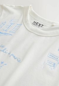 Tricou din bumbac alb, cu desene brodate în albastru ale unor clădiri și text. Guler rotund, mâneci scurte și eticheta "NEXT".