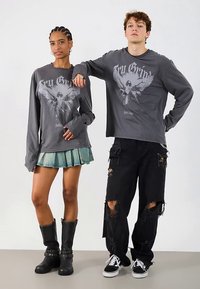 Camisas de manga comprida cinzentas com gráfico de águia; modelo usa uma saia plissada verde e botas pretas até ao joelho, combinadas com calças pretas rasgadas e ténis.