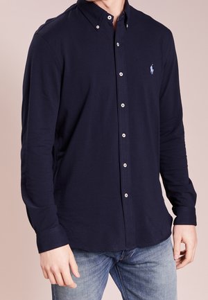 Mand iført en mørkeblå skjorte med knapper med en lille lyseblå broderet logo på brystet og lyseblå jeans.