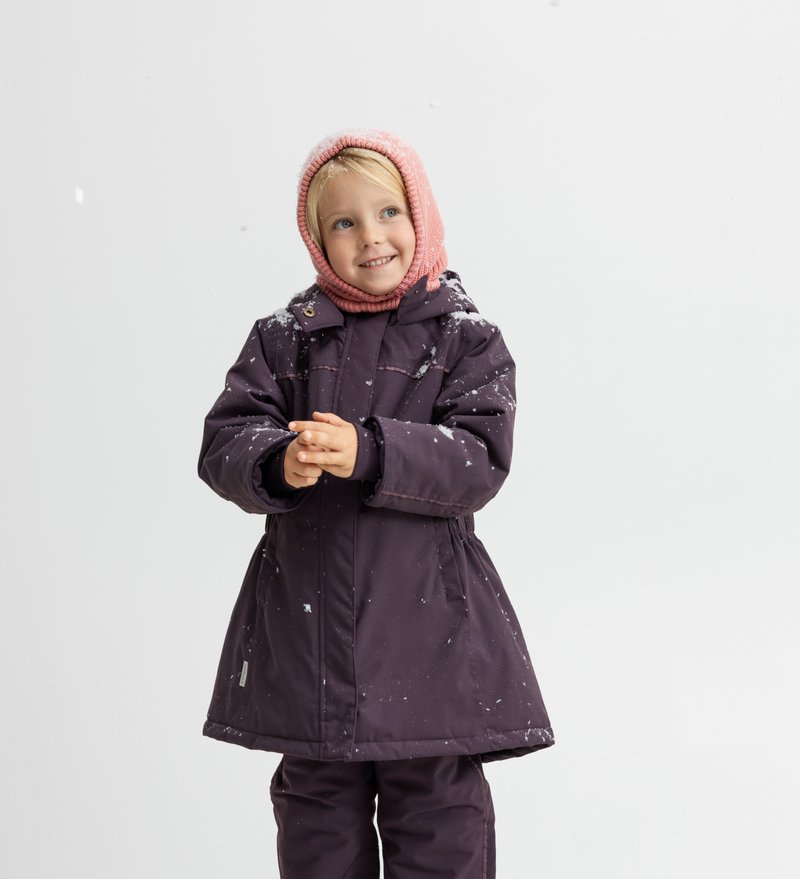 Manteau d'hiver violet avec capuche, doté d'une fermeture éclair et d'une fermeture à bouton, associé à un bonnet en tricot rose clair, de la neige sur le tissu.