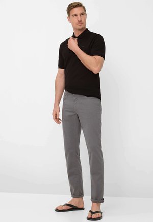 Mann in grauer Hose, schwarzem Poloshirt und schwarzen Flip-Flops, der seinen Kragen richtet und leicht seitlich blickt, vor weißem Hintergrund.