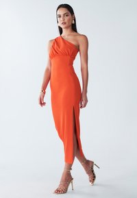 Orange Kleid mit einem Schulterträger, tailliertem Oberteil, seitlichem Schlitz und sanften Drapierungen. Kombiniert mit goldenen Riemchenheeled und minimalem Schmuck.