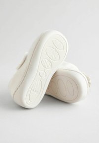 Chaussons blancs en tissu doux avec une semelle en caoutchouc texturée présentant des motifs ovales et un haut du chausson duveteux. Forme arrondie avec un design de talon arrière.