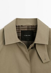 Beige trenchcoat-krave med syninger på slagene, enkelt knap og ternet inderfor med en sort label, der viser "Massimo Dutti".