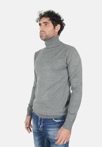 Uomo con capelli ricci scuri che indossa un maglione aderente a collo alto grigio e jeans blu, in piedi e che guarda verso sinistra.