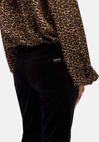 Donna che indossa pantaloni neri e una blusa a maniche lunghe con stampa leopardata, vista di spalle su uno sfondo bianco.