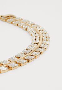 Zlato obarvana ogrlica z izmeničnimi pravokotnimi prozornimi dragulji in gladkimi kovinskimi povezavami, ki prikazuje plastičen dizajn z zanimivo strukturo.