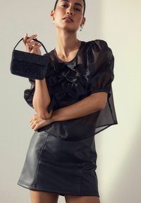 Woman wearing sheer black puff sleeve blouse with bows, black leather skirt, holding a small black handbag with beaded texture.