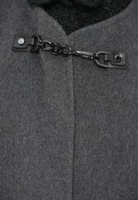 Sivi vuneni kaput s V-izrezom, s crnim kopčanjem od umjetne kože i lancem na ovratniku. Teksturirani materijal, čiste linije.