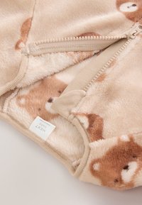 Vêtement pour bébé en polaire beige avec des visages d'ours, partiellement dézippé pour montrer la doublure intérieure douce et pelucheuse ainsi que la protection en tissu de la fermeture éclair.