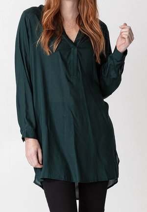 Kobieta z długimi rudymi włosami, ubrana w luźną ciemnozieloną tunikę z długim rękawem i czarne spodnie na jednolitym tle.