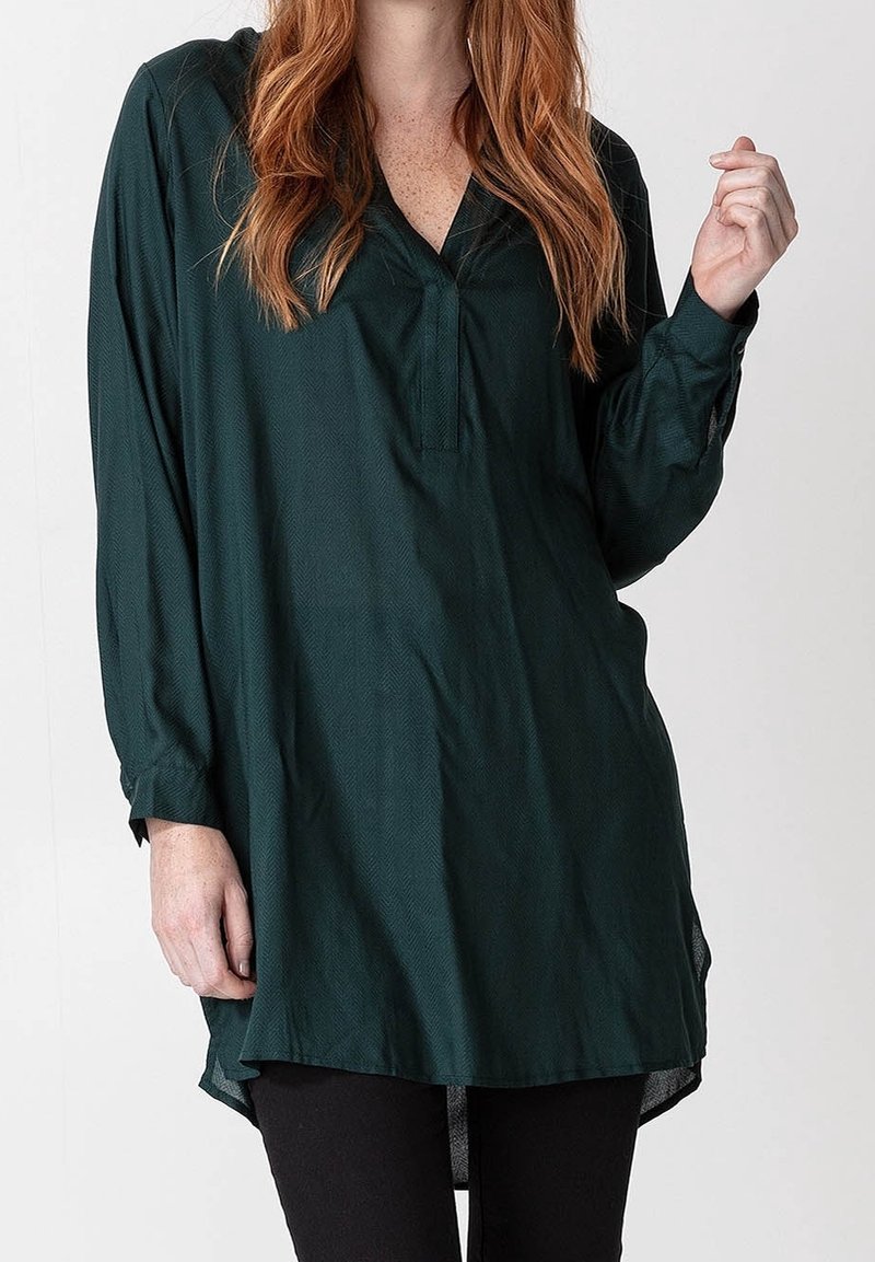 Femme aux longs cheveux roux portant une tunique ample à manches longues vert foncé avec un pantalon noir sur un fond uni.