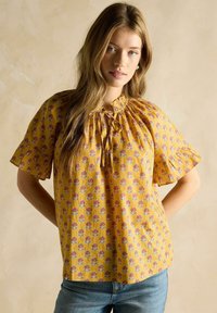 Kollane lilleline pluus lühikeste ripsmete varrukatega, kokku kogutud kaelus ja korduv muster lillade ja punaste lilledega.