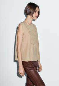 Beige ärmelloses Top mit rundem Ausschnitt, strukturiertem Stoff und Fronttasche. Kombiniert mit braunen Lederhosen, Seitenansicht.