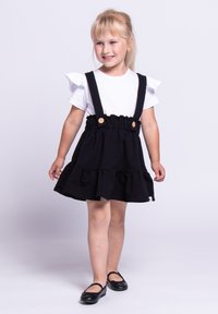 Młoda dziewczynka z blond włosami, ubrana w biały top z falbanami, czarną spódnicę na szelkach i czarne buty Mary Jane, uśmiechająca się i stojąca na białym tle.