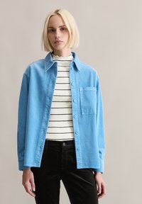 Lichtblauw corduroy overhemd met knoopsluiting aan de voorkant, kraag en een enkele borstzak, gedragen over een wit-zwart gestreept shirt. Zwarte broek.