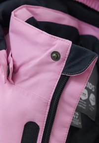 Rosa tyg med en slät yta har en marinblå foder. En silverknapp och en svart dragkedja är synliga. Skötsel etiketter inuti.