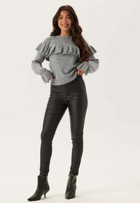Šedý pletený svetr s volantovým výstřihem, doplněný černými kalhotami z umělé kůže a černými kotníkovými botami. Jednoduchý, texturovaný design.