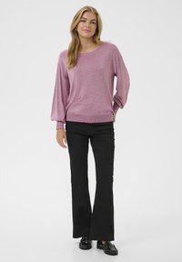 Suéter metálico rosa con mangas largas y voluminosas y puños acanalados. Combinado con pantalones negros de campana y mocasines negros brillantes.