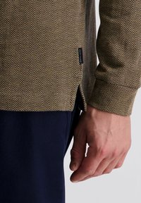 Langsleeve shirt in een visgraatpatroon, met bruine en beige kleuren, met zijsplitsen en een marineblauw textiel onderaan.