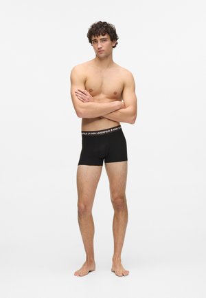Boxers en coton noir avec une ceinture logotée. Les boxers ont une coupe ajustée et une longueur mi-cuisse. Le modèle a la peau et les pieds dénudés.