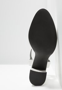 Semelle d'une sandale à talon blanche inclinée vers le haut, montrant une semelle texturée noire et un talon carré sur fond blanc.