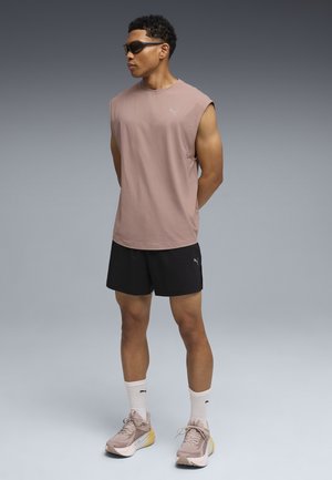 Ljusrosa ärmlös sporttopp med logga, svarta shorts, pastellfärgade sneakers med gula detaljer och vita crew-strumpor, stående pose.