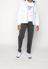 Chaqueta de plumas blanca con capucha, puños elásticos y cremallera frontal, combinada con una sudadera gráfica blanca y jeans gris oscuro. Zapatillas blancas.