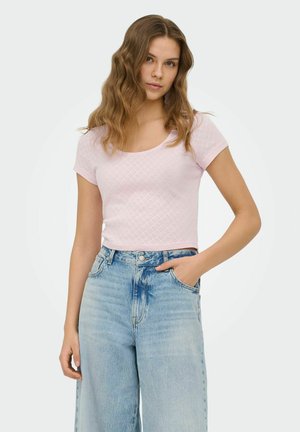 Ung kvinde med bølget hår iført en lyserød kortærmet top og højtaljede lyseblå jeans, stående med den ene hånd i lommen.