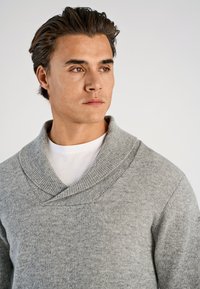 Grå stickad tröja med bred sjalkrage, strukturerat tyg och en figursydd design, lager-på-lager över en vit t-shirt.