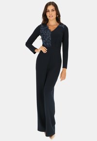 Granatowy kombinezon z długimi rękawami, dekoltem w serek, koronkowym detalem w kwiaty z jednej strony, bezszwowym pasem i szerokimi spodniami. Gładka tekstura materiału.