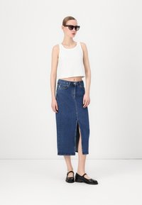 Biely rebrovaný crop top skombinovaný s modrou džínsovou sukňou s predným rozparkom. Model má na sebe čierne topánky a veľké slnečné okuliare.