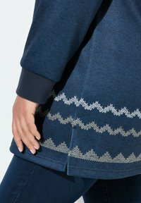 Primo piano di una mano appoggiata vicino all'orlo di un maglione di filato blu con motivi a zigzag bianchi, abbinato a jeans blu scuro.