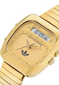 Guld analog-digital klocka med fyrkantig urtavla, texturerat guldarmband, svarta dialaccents och Adidas-logotyp längst ner.