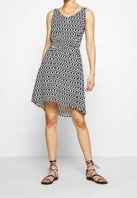 Femme portant une robe sans manches à motifs géométriques noirs et blancs avec un ourlet asymétrique et des sandales plates noires à lacets.