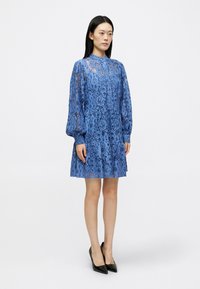 Μπλε κεντητό φόρεμα με μακριά μανίκια και ψηλό λαιμόκοψη, με λουλουδάτα μοτίβα και φουσκωτό στρίφωμα. Φοριέται με μαύρες γωνιώδεις γόβες.