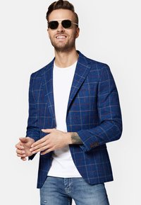 Marynarka w niebieską kratę z czerwonymi i żółtymi liniami, zapinana na pojedynczy guzik, z klapami i akcentami guzikowymi. Nosi się na białej koszulce.