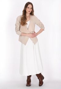 Beige draperet jakke over en hvid kjole med lange ærmer og asymmetrisk kant, parret med brune knæhøje støvler.
