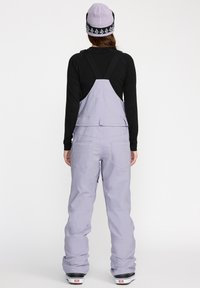 Salopettes de neige techniques violettes avec des bretelles ajustables, deux poches arrière et une coupe décontractée. Associées à un bonnet violet clair.