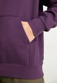 Sweat à capuche violet en mélange de coton, doté d'une poche avant, de poignets et d'un ourlet côtelés, avec des détails de couture visibles.