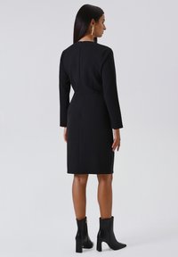 Zwarte langmouw jurk met een getailleerde silhoutte, ronde halslijn en een ritsdetail aan de achterkant. Gemaakt met zwarte enkellaarzen.