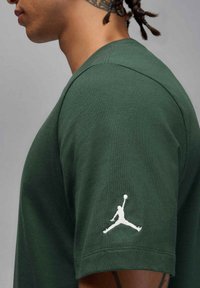 Camiseta de algodón verde oscuro con cuello redondo, costuras visibles y un logo de Jordan bordado en blanco en la manga izquierda.