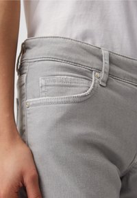 Šedé džíny z denimu s přiléhavým střihem. Obsahují přední kapsu s bílým prošitím a kovovým knoflíkem. Materiál vypadá hladce a je odolný.