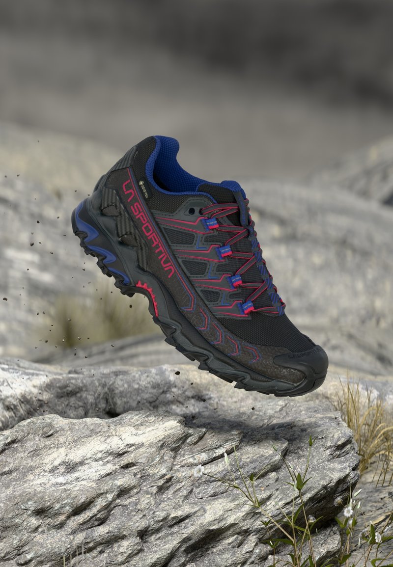 Zapatilla de trail running negra con acentos azules, que presenta una parte superior texturizada, cordones rojos y una suela resistente, suspendida sobre un terreno rocoso.