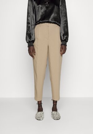 Persoon draagt beige hoge taille taps toelopende broek, zwarte satijnen blouse met lange mouwen en glinsterende geborduurde ballerina's met enkelbandjes, staand tegen een effen achtergrond.