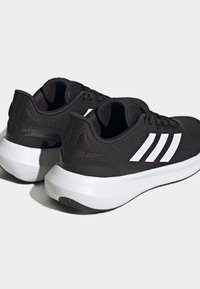 Un par de zapatillas deportivas negras con suelas blancas y tres rayas blancas en los lados, posicionadas ligeramente giradas hacia un lado sobre un fondo claro.