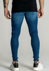 Hoose skinny dalam denim biru sedang dengan pudar halus, menampilkan reka bentuk lima poket klasik dan potongan panjang pergelangan kaki, dipadankan dengan sneakers putih.
